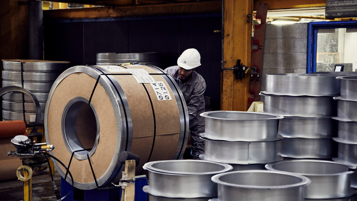 Industrielle Halle mit großen Metallrollen und gestapelten Metallfelgen, Arbeiter mit Schutzhelm im Hintergrund
