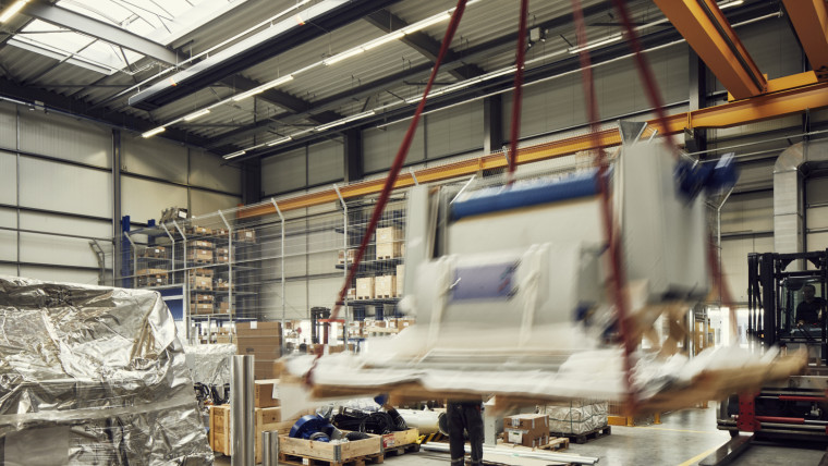 Industrielle Halle mit Hebekran, der eine große Maschine hebt, umgeben von Regalen und verpackten Gütern