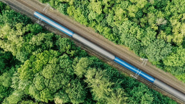 Luftaufnahme eines blauen Zuges auf einer Gleisstrecke, umgeben von dichtem grünem Wald