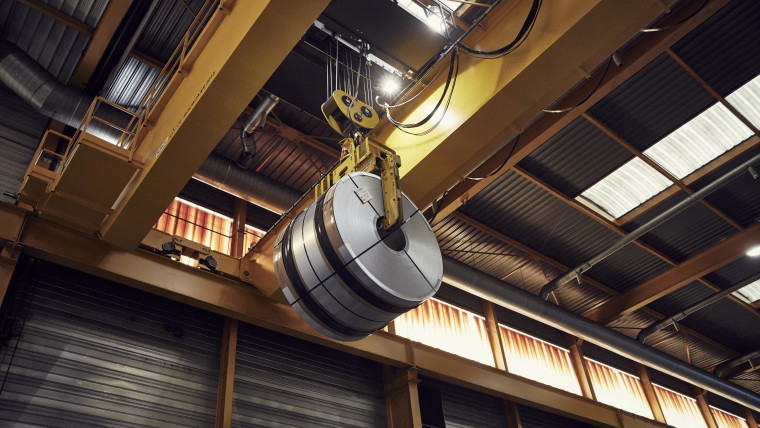 In einer Industriehalle hängt eine große Metallrolle an einem gelben Kranhaken unter der Decke