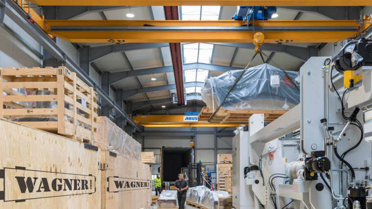 Halle mit großem gelbem ABUS-Kran, der eine mit Folie verpackte Maschine hebt, umgeben von großen Holzkisten mit der Aufschrift WAGNER