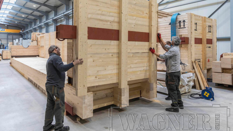 Zwei Männer montieren in einer Halle eine große Holzkiste aus hellen Holzplatten mit roten Metallstreifen